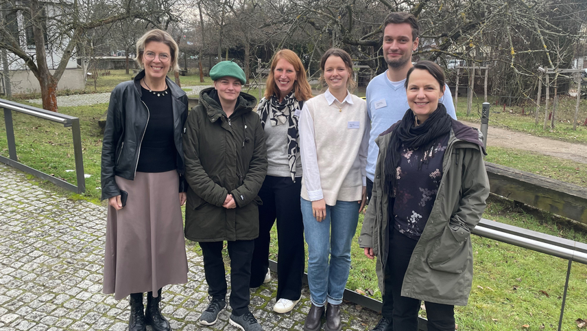 Sechs Personen des LAMBDA-Teams stehen drau&szlig;en nebeneinander und schauen zur Kamera. Sie tragen Jacken und stehen auf einem gepflasterten Weg vor kahlen B&auml;umen.