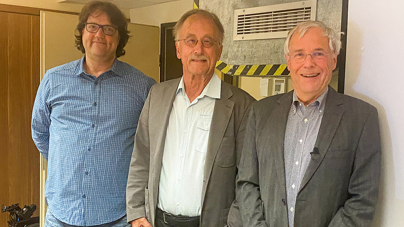 Drei M&auml;nner stehen nebeneinander in einem Raum und l&auml;cheln in die Kamera