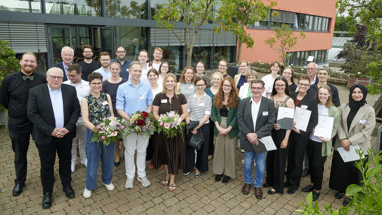 Mehrere Personen lächeln in die Kamera, die Personen in der Mitte halten Blumensträuße