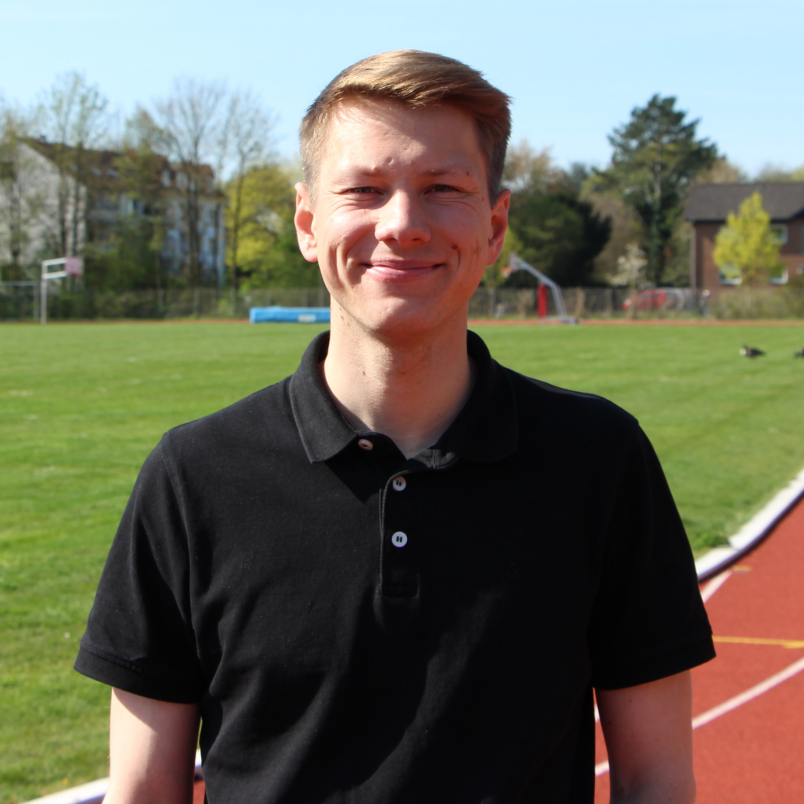 Ein junger Mann l&auml;chelt vor einem Sportplatz.