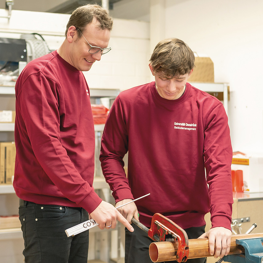 An instructor and an apprentice are standing together at a workbench.