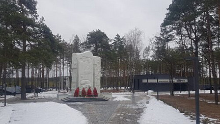 Ein Denkmal aus hellem Stein steht im Zentrum eines gepflasterten Weges, umgeben von schneebedecktem Boden. Vor dem Denkmal sind rote Blumengestecke platziert. Die Umgebung besteht aus hohen, schlanken Bäumen, die einen Wald bilden. Rechts vom Denkmal befindet sich ein dunkles, modernes Gebäude mit großen Fenstern. Ein bewölkter Himmel ist im Hintergrund zu sehen.