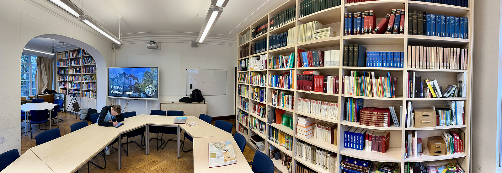 Ein heller Raum mit mehreren Tischen, die in U-Form angeordnet sind, und St&uuml;hlen darum herum. Eine Person sitzt an einem Tisch und blickt auf ein Ger&auml;t vor sich.  An der rechten Wand stehen gro&szlig;e Regale, die vollst&auml;ndig mit B&uuml;chern gef&uuml;llt sind. Im hinteren Bereich befindet sich ein Bildschirm, auf dem &bdquo;Herzlich Willkommen!&ldquo; zu lesen ist.  Links f&uuml;hrt ein Durchgang in einen weiteren Raum mit weiteren Regalen und einem Fenster. An der Decke h&auml;ngen l&auml;ngliche Lampen.