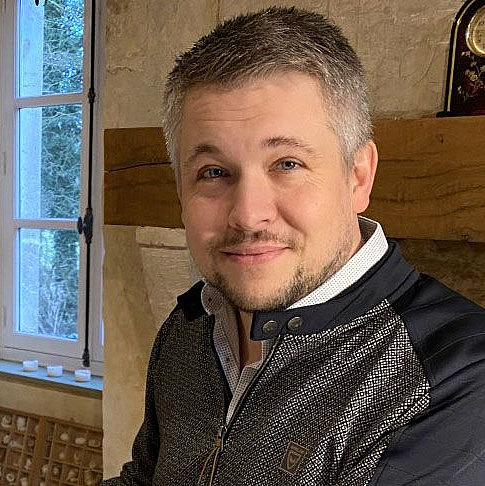 Ein Mann mit kurzem, graumeliertem Haar und einem leichten Lächeln steht in einem rustikalen Raum. Er trägt eine Jacke mit kontrastierenden Kragen und Akzenten. Im Hintergrund sind ein Fenster mit Blick ins Freie und eine Steinwand zu sehen