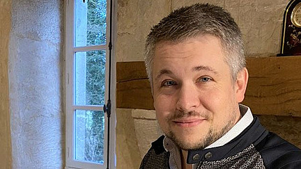 Ein Mann mit kurzem, graumeliertem Haar und einem leichten Lächeln steht in einem rustikalen Raum. Er trägt eine Jacke mit kontrastierenden Kragen und Akzenten. Im Hintergrund sind ein Fenster mit Blick ins Freie und eine Steinwand zu sehen