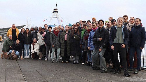 A group of 40 to 50 people smile for the camera, with ships and water visible in the background.