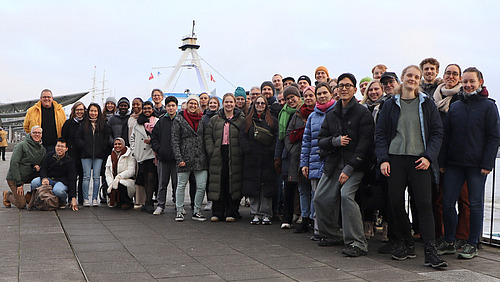 A group of 40 to 50 people smile for the camera, with ships and water visible in the background.