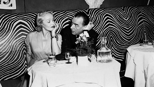 A black and white photo shows Marlene Dietrich and Erich Maria Remarque sitting in a bar, engrossed in conversation.