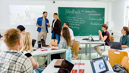 Lernende an Gruppentischen schauen in Richtung Tafel und Lehrende Personen