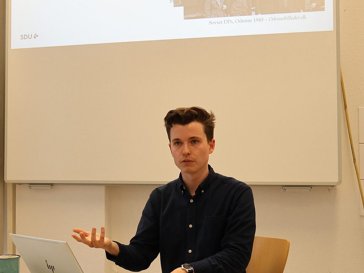 Das Foto zeigt eine Person, die an einem Tisch sitzt und zu einem Vortrag oder einer Präsentation spricht. Im Hintergrund wird eine Folie projiziert mit der Überschrift "Context 3: Who were the DPs in Denmark?". Die Präsentation enthält Text und ein historisches Foto. Auf dem Tisch vor der Person liegt ein Laptop, daneben sind Notizen und ein Becher sichtbar.