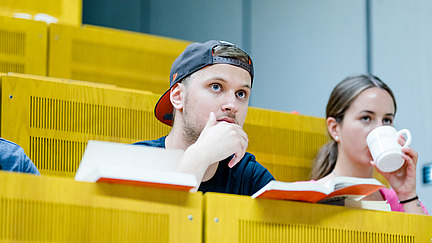Zwei Studierende auf Sitzplätzen in einem Hörsaal. Eine von ihnen trinkt etwas aus einer Tasse.