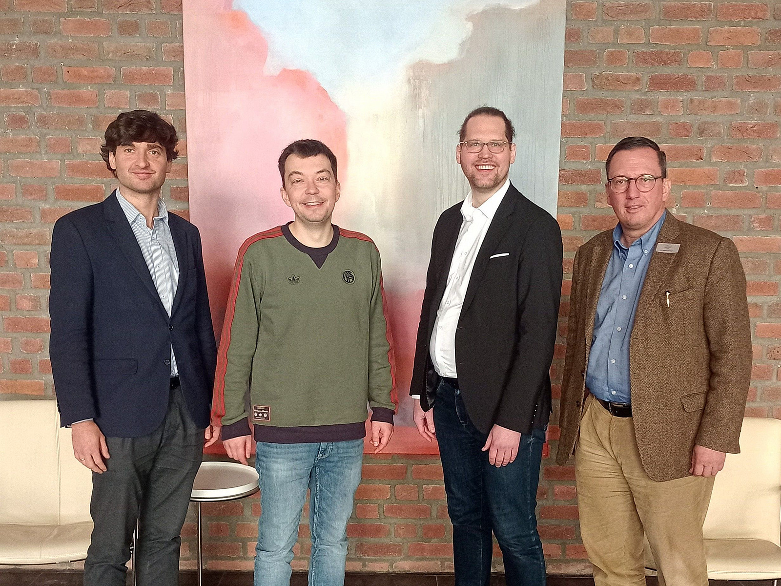Vier Personen stehen nebeneinander vor einer Wand aus roten Ziegelsteinen. Hinter ihnen h&auml;ngt ein gro&szlig;es Bild mit hellen und dunklen Farbfl&auml;chen.  Die Personen sind in einer Reihe angeordnet und schauen in Richtung Kamera. Sie tragen unterschiedliche Kleidung, darunter Jacken, Hemden und ein Pullover.  Im Vordergrund ist ein Teil eines Tisches oder einer Sitzgelegenheit zu sehen.