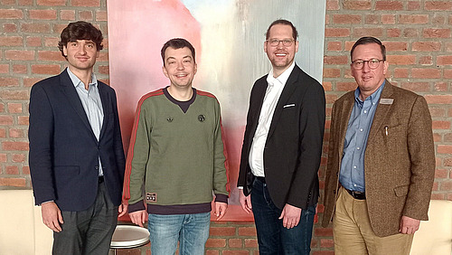 Vier Personen stehen nebeneinander vor einer Wand aus roten Ziegelsteinen. Hinter ihnen h&auml;ngt ein gro&szlig;es Bild mit hellen und dunklen Farbfl&auml;chen.  Die Personen sind in einer Reihe angeordnet und schauen in Richtung Kamera. Sie tragen unterschiedliche Kleidung, darunter Jacken, Hemden und ein Pullover.  Im Vordergrund ist ein Teil eines Tisches oder einer Sitzgelegenheit zu sehen.