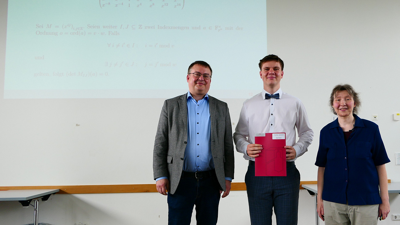 Ein Student mit Studiendekanin und Dekan stehen nebeneinander. Der Student hat eine Mappe in der Hand