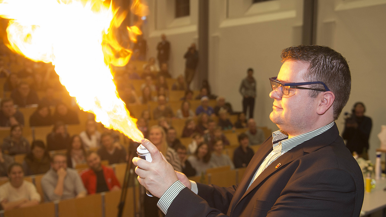 Ein Mann lässt eine Flamme aufflammen, im Hintergrund sind Personen in einem Hörsaal zu sehen.