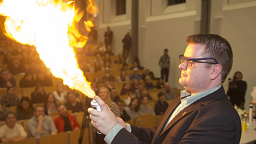 Ein Mann lässt eine Flamme aufflammen, im Hintergrund sind Personen in einem Hörsaal zu sehen.