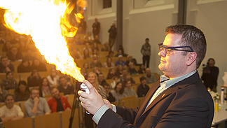 A man lights a flame; people can be seen in a lecture hall in the background.