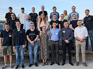 21 Personen haben sich in drei Reihen zu einem Gruppenbild aufgestellt. Im Hintergrund ein großes Gebäude.