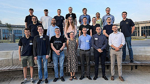 21 Personen haben sich in drei Reihen zu einem Gruppenbild aufgestellt. Im Hintergrund ein großes Gebäude.