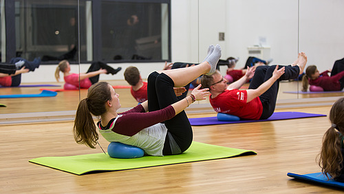 A group of people participates in a Pilates class, performing exercises on mats. The focus is on strengthening the core and improving flexibility.