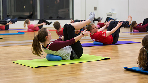 Eine Gruppe von Menschen nimmt an einem Pilates-Kurs teil und führt Übungen auf Matten aus. Der Fokus liegt auf der Stärkung der Körpermitte und der Verbesserung der Flexibilität.