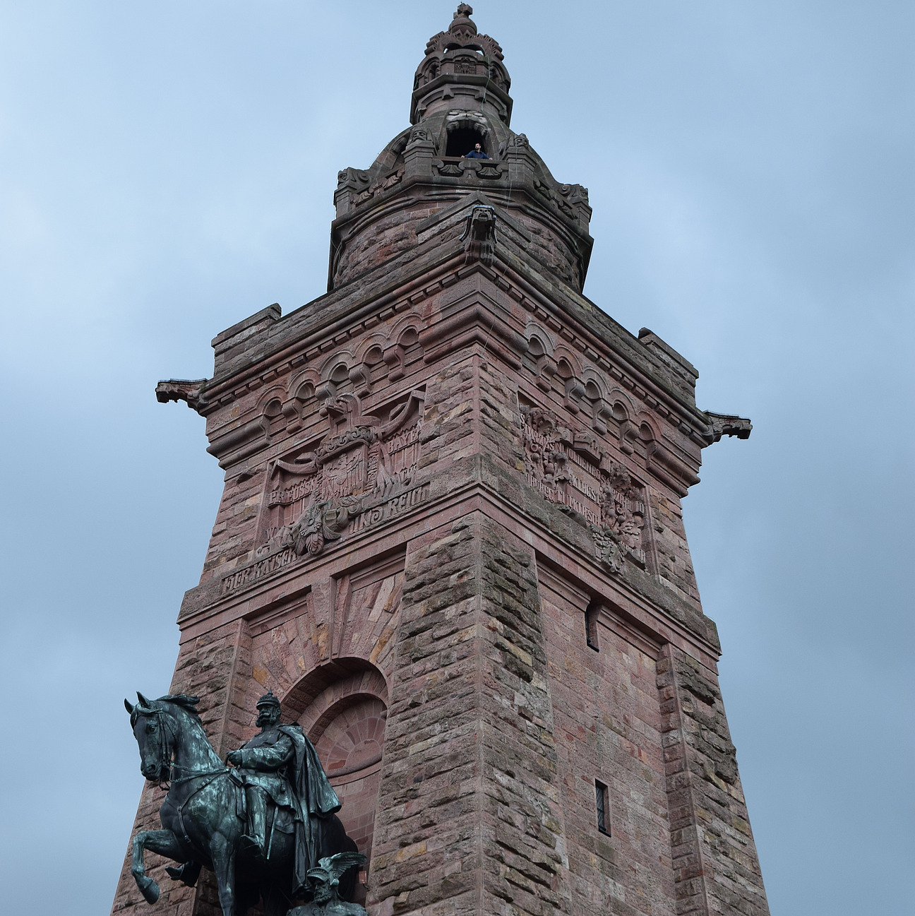 Das Bild zeigt ein großes Denkmal. Es ist eine hohe rötliche Säule und davor steht eine Reiterstandbild.