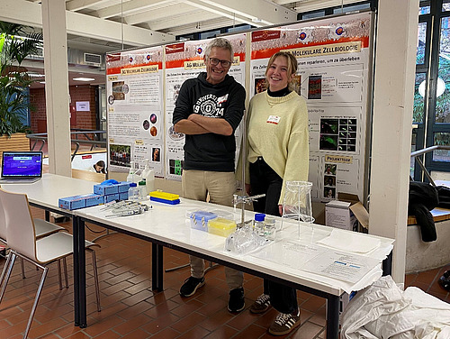 A man and a woman are standing behind a table with various laboratory items on it, smiling.