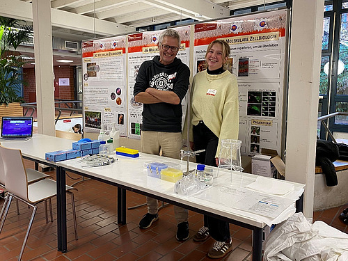 Ein Mann und eine Frau stehen lächelnd hinter einem Tisch, auf dem sich unterschiedliche Labor-Gegenstände befinden