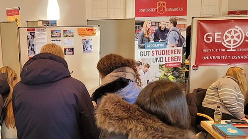 Pupils stand crowded together, with posters and information material bearing the Osnabrück University logo visible in the background.