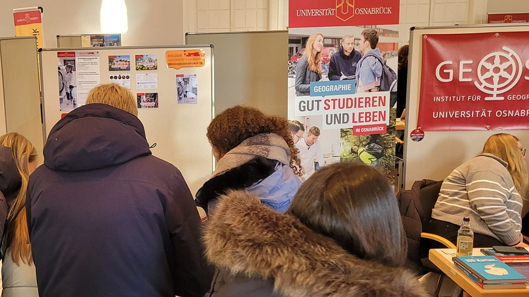 Pupils stand crowded together, with posters and information material bearing the Osnabrück University logo visible in the background.