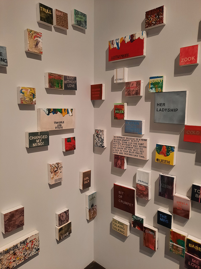 Das Bild zeigt eine Ausstellung mit vielen kleinen, individuell gestalteten Leinwänden, die mit Texten und farbigen Mustern an den Wänden eines Raums angebracht sind.