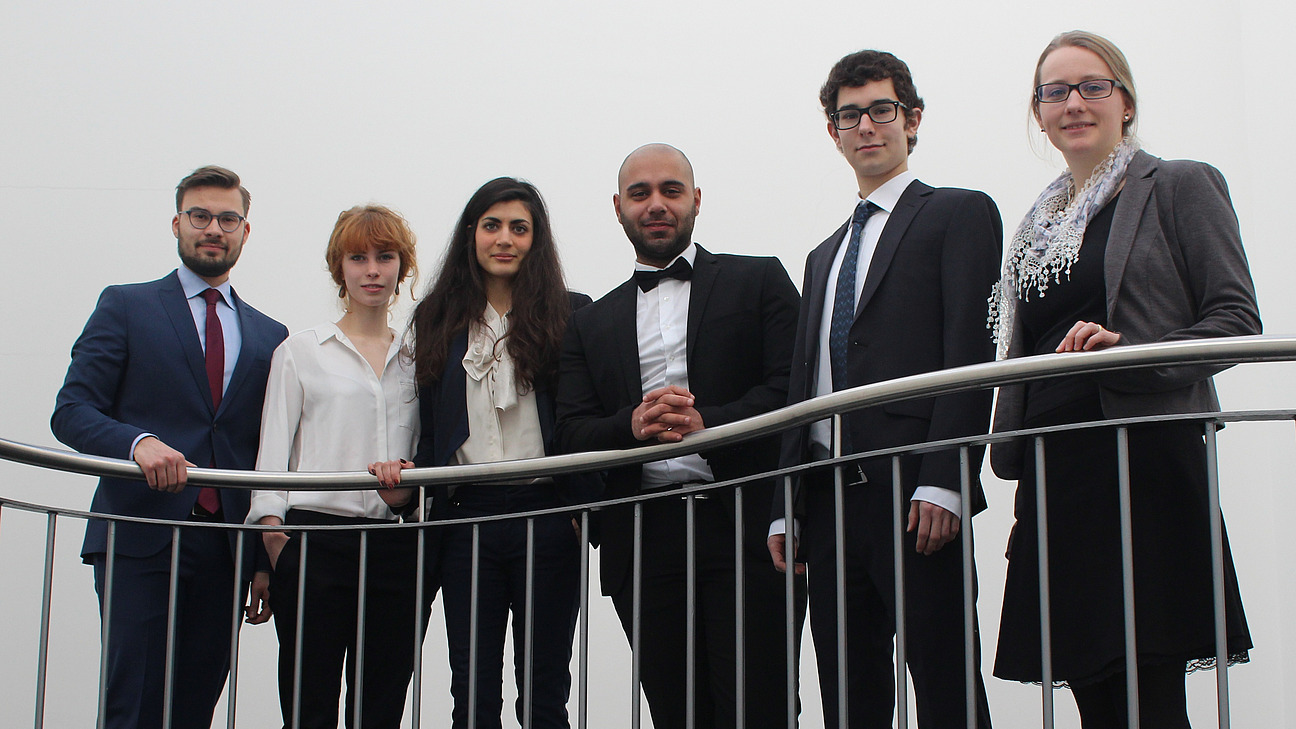 Drei M&auml;nner und drei Frauen in einer Reihe hinter halbhohem Metallgel&auml;nder stehend