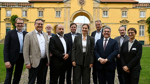 Eine Gruppe von Personen in förmlicher Kleidung steht vor einem historischen Torbogen. Alle lächeln in die Kamera.