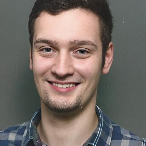 Porträt eines jungen Mannes mit kurzem dunklem Haar und kurzem Bart. Er trägt ein kariertes Hemd und lächelt in die Kamera.