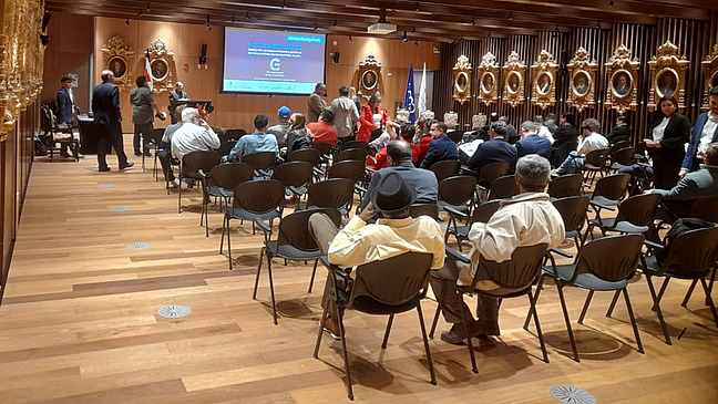 Ein gro&szlig;er festlicher Saal, gef&uuml;llt mit Menschen, die auf St&uuml;hlen sitzen. Auf einer Leinwand an der Vorderseite des Raums ist eine Pr&auml;sentation zu sehen. Eine Person steht und spricht, wahrscheinlich h&auml;lt sie eine Pr&auml;sentation.