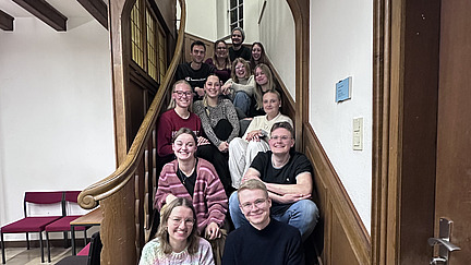 Eine Gruppe junger Erwachsener sitzt und steht gemeinsam auf einer geschwungenen Holztreppe in einem Geb&auml;ude. Die Personen sind dicht beieinander angeordnet und schauen in Richtung Kamera. Viele l&auml;cheln, die Stimmung wirkt freundlich und entspannt.  Die Treppe hat ein geschwungenes Gel&auml;nder aus Holz und wird von einem gro&szlig;en Rundbogen eingerahmt. Im Hintergrund sind helle W&auml;nde und ein Fenster zu sehen. Die Gruppe scheint sich f&uuml;r ein gemeinsames Foto versammelt zu haben.