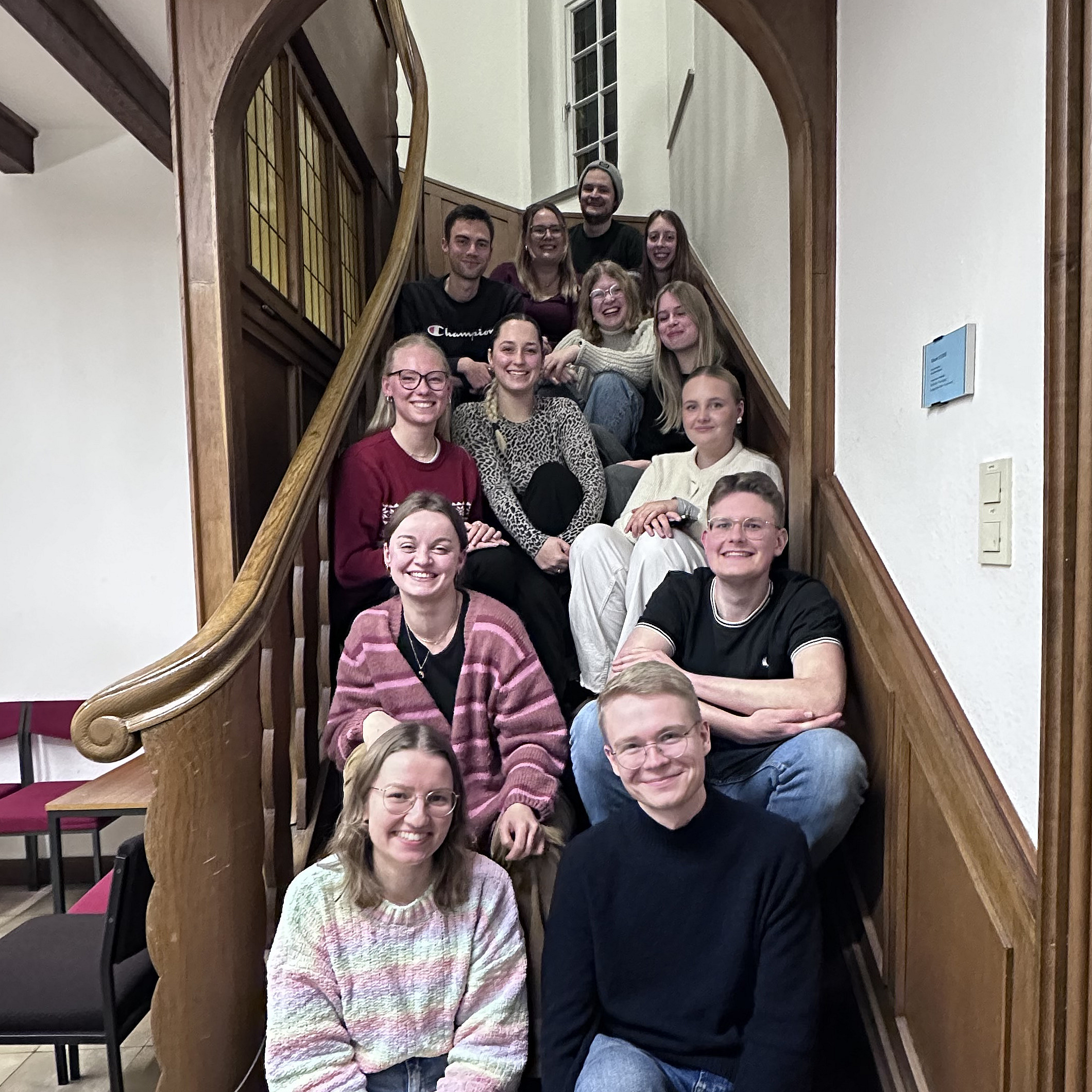 Eine Gruppe junger Erwachsener sitzt und steht gemeinsam auf einer geschwungenen Holztreppe in einem Geb&auml;ude. Die Personen sind dicht beieinander angeordnet und schauen in Richtung Kamera. Viele l&auml;cheln, die Stimmung wirkt freundlich und entspannt.  Die Treppe hat ein geschwungenes Gel&auml;nder aus Holz und wird von einem gro&szlig;en Rundbogen eingerahmt. Im Hintergrund sind helle W&auml;nde und ein Fenster zu sehen. Die Gruppe scheint sich f&uuml;r ein gemeinsames Foto versammelt zu haben.