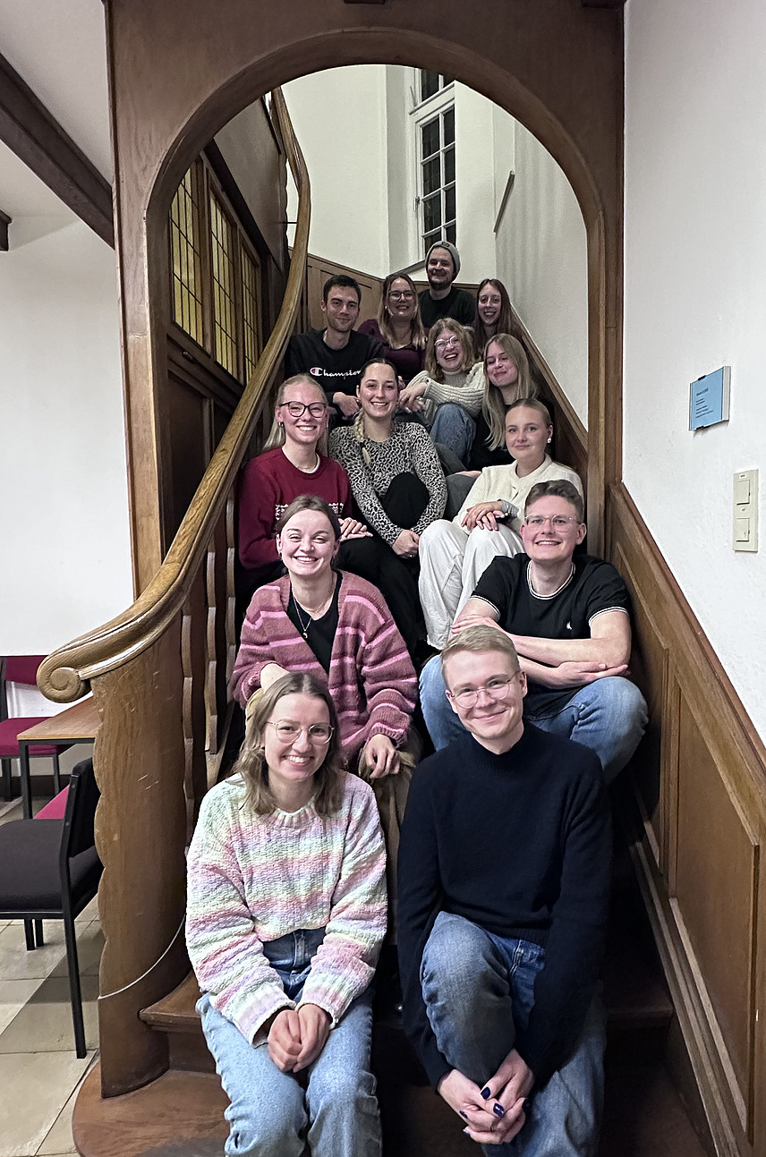 Eine Gruppe junger Erwachsener sitzt und steht gemeinsam auf einer geschwungenen Holztreppe in einem Geb&auml;ude. Die Personen sind dicht beieinander angeordnet und schauen in Richtung Kamera. Viele l&auml;cheln, die Stimmung wirkt freundlich und entspannt.  Die Treppe hat ein geschwungenes Gel&auml;nder aus Holz und wird von einem gro&szlig;en Rundbogen eingerahmt. Im Hintergrund sind helle W&auml;nde und ein Fenster zu sehen. Die Gruppe scheint sich f&uuml;r ein gemeinsames Foto versammelt zu haben.