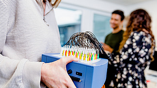 An EEG device is being held by a young woman. In the background, a young man and another young woman can be seen.