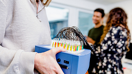 An EEG device is being held by a young woman. In the background, a young man and another young woman can be seen.