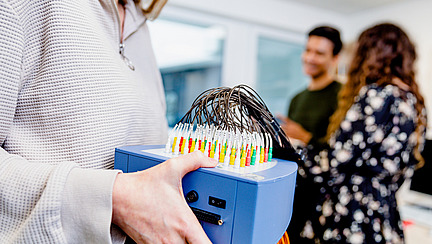 An EEG device is being held by a young woman. In the background, a young man and another young woman can be seen.