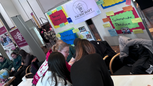 Mehrere Studierende sitzen eng beieinander an einem Tisch und arbeiten gemeinsam an Aufgaben. Sie beugen sich &uuml;ber Materialien und scheinen sich auszutauschen.  Im Hintergrund ist eine Wand mit vielen bunten Zetteln und Plakaten zu sehen. Auf einem gr&ouml;&szlig;eren Plakat steht &bdquo;Fachschaft Katholische Theologie&ldquo;. Die farbigen Zettel enthalten kurze Begriffe und Themen.   Weitere Personen sitzen im Hintergrund oder bewegen sich durch den Raum. Die Atmosph&auml;re ist lebendig und gemeinschaftlich.
