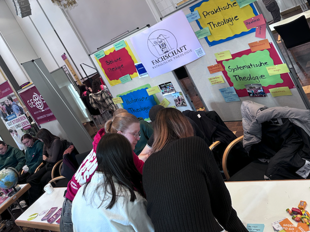 Mehrere Studierende sitzen eng beieinander an einem Tisch und arbeiten gemeinsam an Aufgaben. Sie beugen sich &uuml;ber Materialien und scheinen sich auszutauschen.  Im Hintergrund ist eine Wand mit vielen bunten Zetteln und Plakaten zu sehen. Auf einem gr&ouml;&szlig;eren Plakat steht &bdquo;Fachschaft Katholische Theologie&ldquo;. Die farbigen Zettel enthalten kurze Begriffe und Themen.   Weitere Personen sitzen im Hintergrund oder bewegen sich durch den Raum. Die Atmosph&auml;re ist lebendig und gemeinschaftlich.