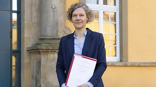 Frau im Anzug steht auf der Treppe vor dem Eingang eines gro&szlig;en Geb&auml;udes und schaut in die Kamera