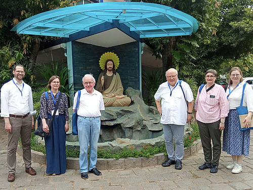 Sechs Personen stehen nebeneinander vor einer kleinen Anlage mit Steinen und Pflanzen. Hinter ihnen befindet sich eine sitzende Figur auf einem Felsen, umgeben von einem runden Hintergrund.  &Uuml;ber der Szene ist eine Konstruktion aus Glas oder transparentem Material mit blauen Tr&auml;gern angebracht. Die Umgebung ist begr&uuml;nt mit B&auml;umen und Str&auml;uchern.  Die Personen stehen in einer Reihe, mit etwas Abstand zueinander, und schauen in Richtung Kamera.