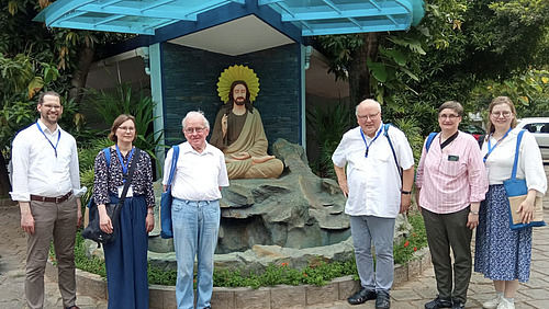 Sechs Personen stehen nebeneinander vor einer kleinen Anlage mit Steinen und Pflanzen. Hinter ihnen befindet sich eine sitzende Figur auf einem Felsen, umgeben von einem runden Hintergrund.  &Uuml;ber der Szene ist eine Konstruktion aus Glas oder transparentem Material mit blauen Tr&auml;gern angebracht. Die Umgebung ist begr&uuml;nt mit B&auml;umen und Str&auml;uchern.  Die Personen stehen in einer Reihe, mit etwas Abstand zueinander, und schauen in Richtung Kamera.