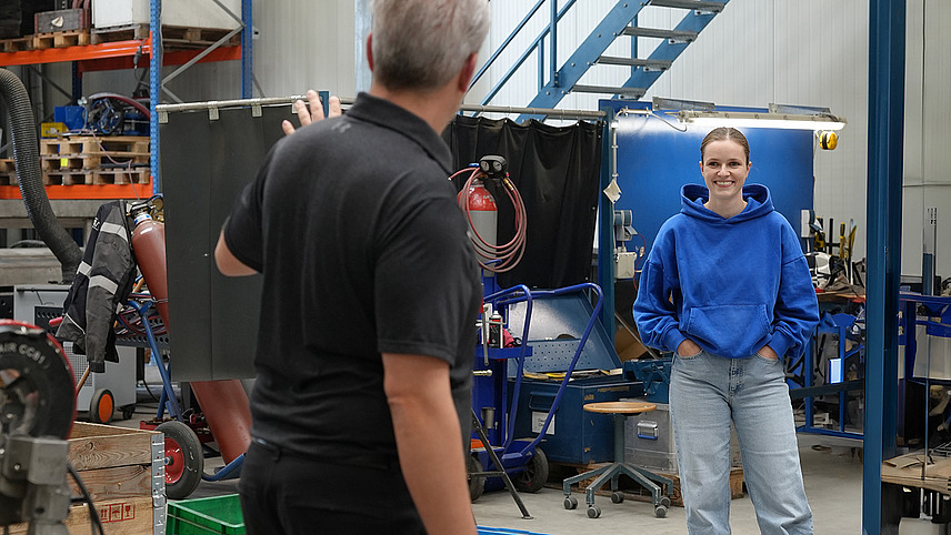 Zwei Personen stehen in einem Werkstattbereich mit technischer Ausr&uuml;stung und scheinen ein Projekt zu besprechen oder zu erkl&auml;ren. Im Hintergrund sind verschiedene Ger&auml;te und Materialien zu sehen.