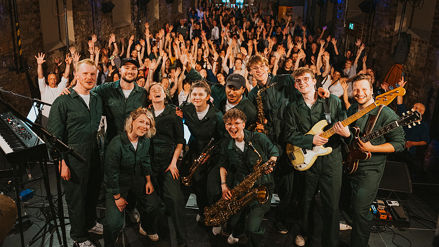 Eine Band steht auf einer B&uuml;hne vor einem Publikum. Alle jubeln in die Kamera