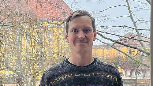 Dr. Benjamin B&uuml;scher steht in einem Pullover vor dem Schloss der Universit&auml;t Osnabr&uuml;ck.