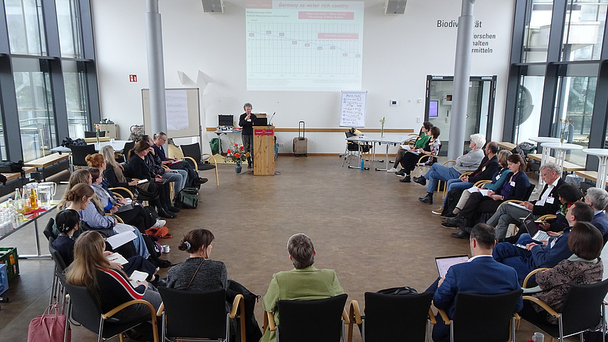 A group of 24 people is sitting in a semicircle in a spacious, bright room with big windows, listening to a woman standing in front who is giving a talk